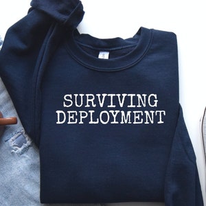 May include: A navy blue sweatshirt with the text "SURVIVING DEPLOYMENT" in white lettering. The sweatshirt is laid flat on a white surface with a pair of gray sneakers and blue jeans.