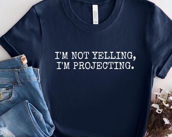 May include: Navy blue t-shirt with the text "I'M NOT YELLING, I'M PROJECTING." The shirt is paired with blue denim jeans. A small bouquet of white and yellow flowers is placed near the shirt.