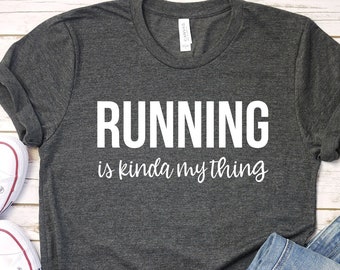 May include: A dark gray t-shirt with the text "RUNNING is kinda my thing" in white lettering. The shirt is displayed on a wooden surface with a pair of white sneakers and blue jeans.
