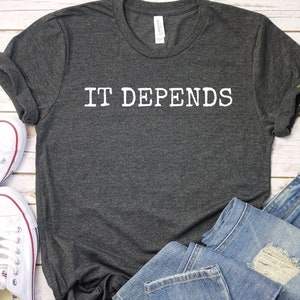 May include: A dark grey t-shirt with the text "IT DEPENDS" printed in white on the front. The shirt is laid out on a white wooden surface with a pair of white trainers and a pair of blue jeans.