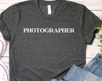May include: A dark gray t-shirt with the word "PHOTOGRAPHER" in white block letters. The shirt is paired with light blue denim jeans and white canvas sneakers with red accents.