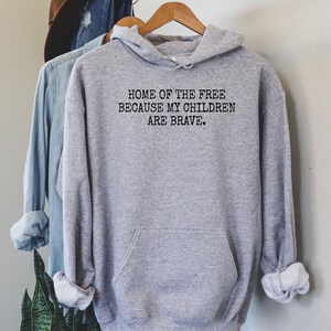 May include: A heather gray hoodie with the text "HOME OF THE FREE BECAUSE MY CHILDREN ARE BRAVE" in black. The hoodie has a front pocket and is hanging on a wooden hanger. Other clothing items and a hat are visible in the background.