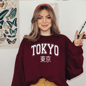 May include: A maroon sweatshirt with the word "TOKYO" in white letters and the Japanese characters for "Tokyo" underneath. The model is wearing the sweatshirt and has long brown hair.