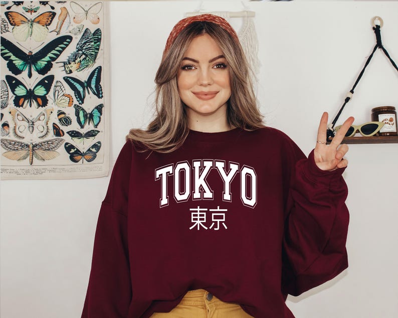 May include: A maroon sweatshirt with the word "TOKYO" in white letters and the Japanese characters for "Tokyo" underneath. The model is wearing the sweatshirt and has long brown hair.
