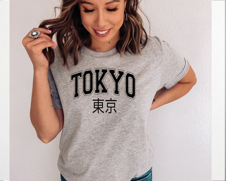 K&ouml;nnte beinhalten: Ein graues T-Shirt mit dem Wort "TOKYO" in schwarzen Blockbuchstaben und den japanischen Schriftzeichen f&uuml;r "Tokyo" darunter.