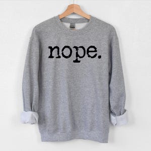 May include: A heather gray sweatshirt with the word "nope" printed in black, displayed on a wooden hanger. The long sleeves are cuffed at the wrists. The sweatshirt is made of a soft material.