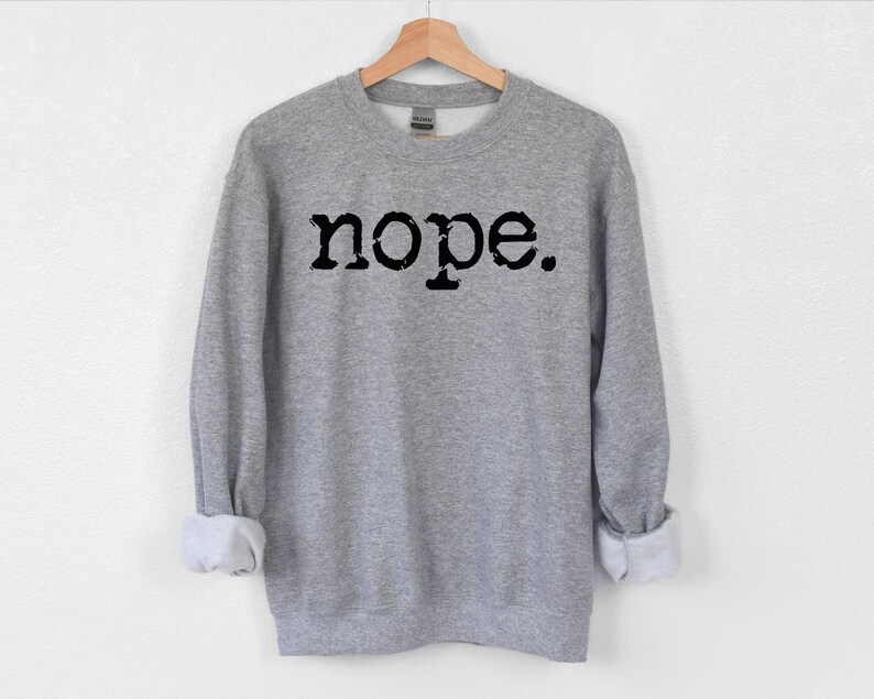 May include: A heather gray sweatshirt with the word "nope" printed in black, displayed on a wooden hanger. The long sleeves are cuffed at the wrists. The sweatshirt is made of a soft material.
