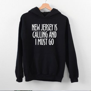 May include: Black hooded sweatshirt with the text "NEW JERSEY IS CALLING AND I MUST GO" in white. The sweatshirt has a front pocket and a drawstring hood. The garment is hanging on a wooden hanger.