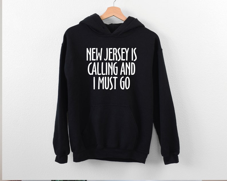 May include: Black hooded sweatshirt with the text "NEW JERSEY IS CALLING AND I MUST GO" in white. The sweatshirt has a front pocket and a drawstring hood. The garment is hanging on a wooden hanger.