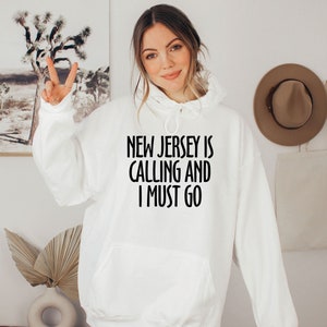 May include: White hoodie with the text "NEW JERSEY IS CALLING AND I MUST GO" in black. The hoodie has a front pocket and a drawstring hood. The person is wearing the hoodie and giving a peace sign.