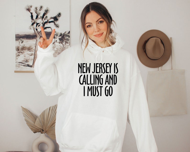 May include: White hoodie with the text "NEW JERSEY IS CALLING AND I MUST GO" in black. The hoodie has a front pocket and a drawstring hood. The person is wearing the hoodie and giving a peace sign.