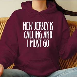 May include: A burgundy hoodie featuring the white text "NEW JERSEY IS CALLING AND I MUST GO". The hoodie has a hood and a front pocket. The text is in a bold, sans-serif font.