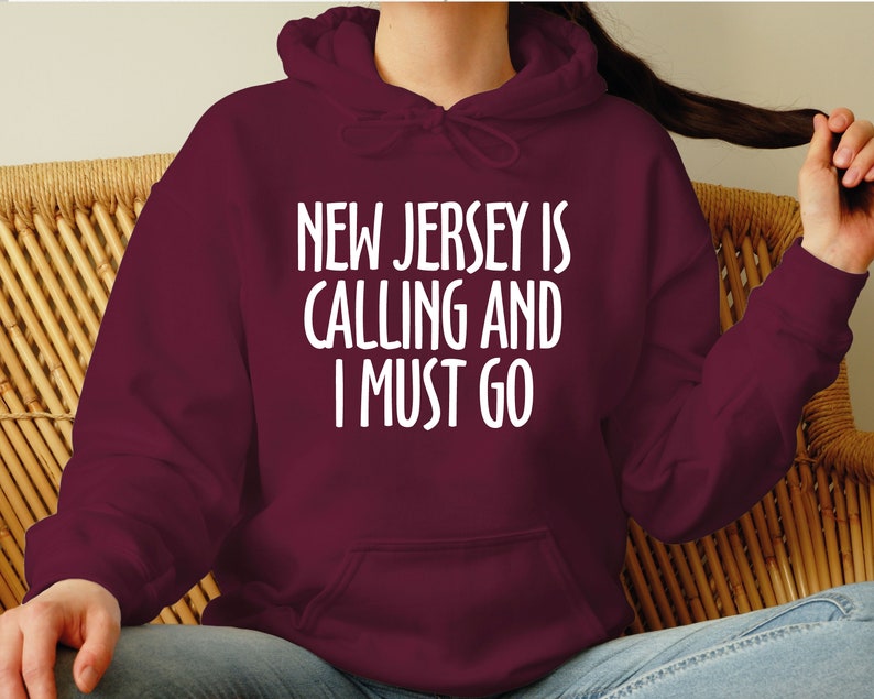 May include: A burgundy hoodie featuring the white text "NEW JERSEY IS CALLING AND I MUST GO". The hoodie has a hood and a front pocket. The text is in a bold, sans-serif font.