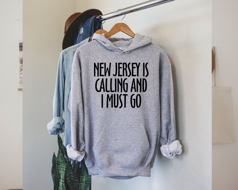 May include: A grey hoodie with the black text "NEW JERSEY IS CALLING AND I MUST GO". The hoodie has a hood and a front pocket. Other clothing and a hat are visible in the background, hanging on a rail.