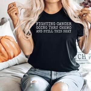 Op de afbeelding: Zwart T-shirt met witte tekst die "Fighting Cancer Going Thru Chemo And Still This Sexy" zegt