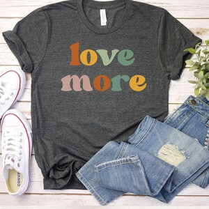 May include: Dark gray t-shirt with the words "love more" in colorful retro font. The shirt is paired with blue jeans and white canvas sneakers. The image is set on a white wooden surface.