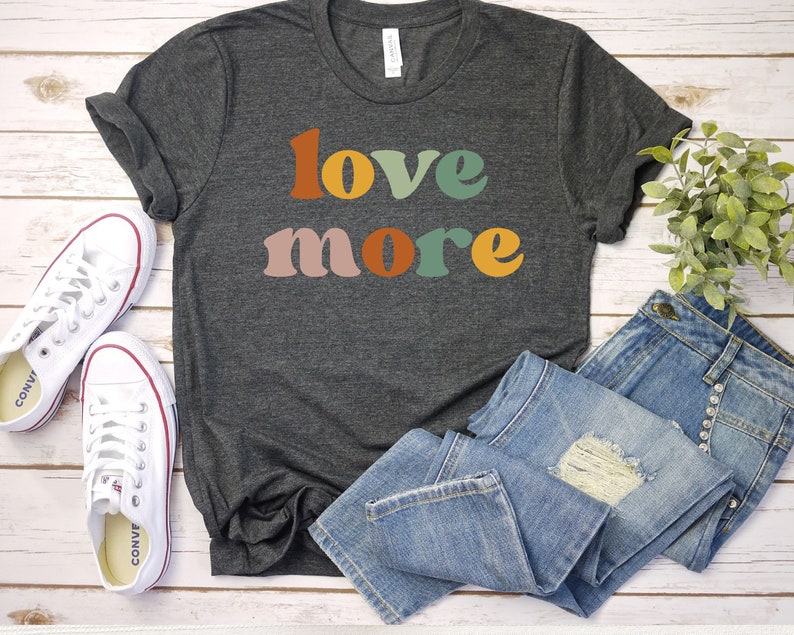 May include: Dark gray t-shirt with the words "love more" in colorful retro font. The shirt is paired with blue jeans and white canvas sneakers. The image is set on a white wooden surface.