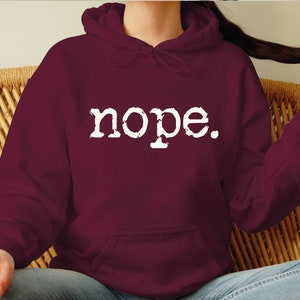 May include: A maroon hoodie with the word "nope" printed in white, featuring a drawstring hood and a front pocket. The hoodie is made of a soft material and is suitable for casual wear.