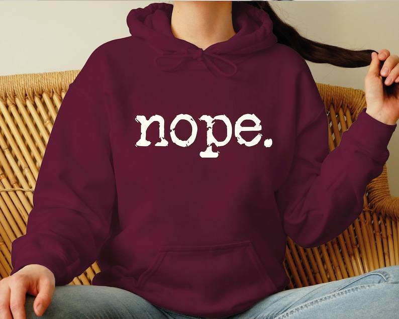 May include: A maroon hoodie with the word "nope" printed in white, featuring a drawstring hood and a front pocket. The hoodie is made of a soft material and is suitable for casual wear.