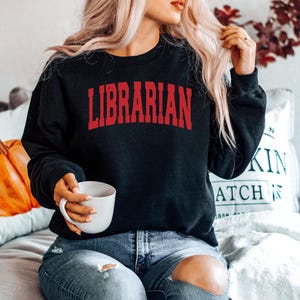 May include: A woman wearing a black sweatshirt with the word "LIBRARIAN" in red letters. She is sitting on a couch and holding a white mug.