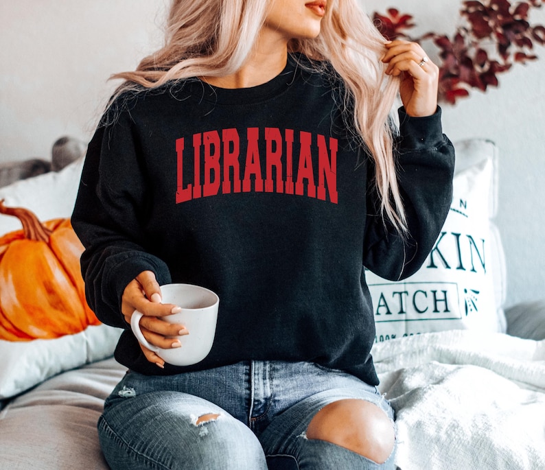 May include: A woman wearing a black sweatshirt with the word "LIBRARIAN" in red letters. She is sitting on a couch and holding a white mug.