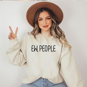 Może przedstawiać: Kobieta w jasnobeżowej bluzie z nadrukiem "EW, PEOPLE" w kolorze czarnym. Ma na sobie brązowy kapelusz i długie brązowe włosy.