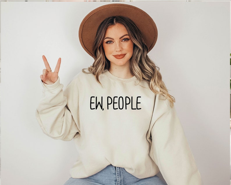 Może przedstawiać: Kobieta w jasnobeżowej bluzie z nadrukiem "EW, PEOPLE" w kolorze czarnym. Ma na sobie brązowy kapelusz i długie brązowe włosy.