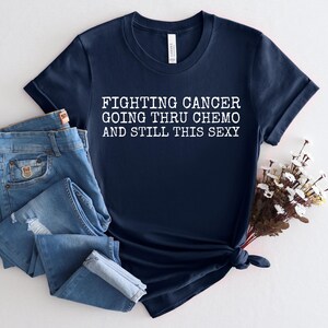 Op de afbeelding: Donkerblauw T-shirt met witte tekst die "Fighting Cancer Going Thru Chemo And Still This Sexy" luidt.