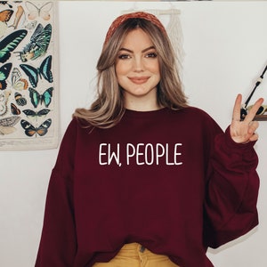 Może przedstawiać: Kobieta w bordowej bluzie z napisem "EW, PEOPLE" w białych literach. Uśmiecha się i pokazuje znak pokoju prawą ręką.