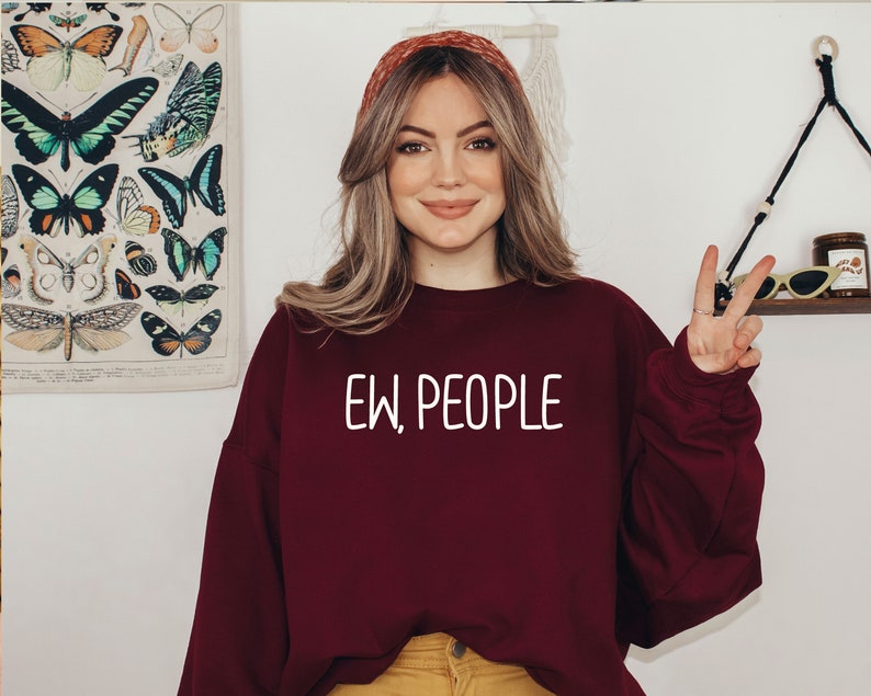 Może przedstawiać: Kobieta w bordowej bluzie z napisem "EW, PEOPLE" w białych literach. Uśmiecha się i pokazuje znak pokoju prawą ręką.