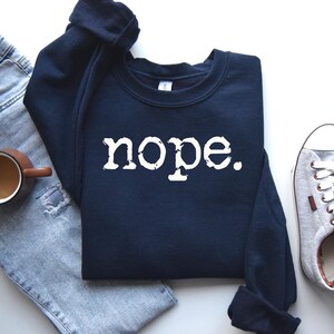 May include: A navy blue sweatshirt with the word "nope" in white, block letters. The sweatshirt is laid flat next to a pair of light blue jeans, a brown mug of coffee, and a pair of gray sneakers.