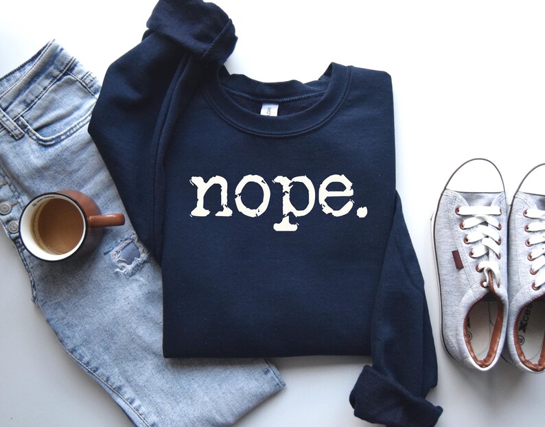 May include: A navy blue sweatshirt with the word "nope" in white, block letters. The sweatshirt is laid flat next to a pair of light blue jeans, a brown mug of coffee, and a pair of gray sneakers.