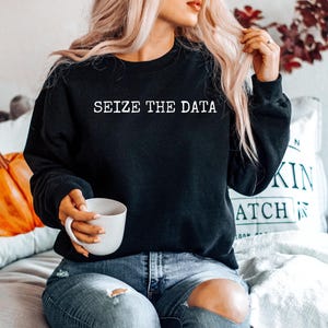 K&ouml;nnte beinhalten: Ein schwarzer Pullover mit dem Text "Seize the Data" in wei&szlig;en Buchstaben. Das Model tr&auml;gt den Pullover mit einer blauen Jeans und h&auml;lt eine wei&szlig;e Tasse.