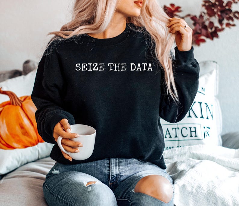 K&ouml;nnte beinhalten: Ein schwarzer Pullover mit dem Text "Seize the Data" in wei&szlig;en Buchstaben. Das Model tr&auml;gt den Pullover mit einer blauen Jeans und h&auml;lt eine wei&szlig;e Tasse.