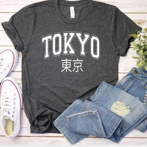 K&ouml;nnte beinhalten: Ein dunkelgraues T-Shirt mit wei&szlig;em Text, auf dem "TOKYO" und die japanischen Schriftzeichen f&uuml;r Tokio stehen. Das Shirt liegt flach auf einer Holzoberfl&auml;che mit einem Paar wei&szlig;en Turnschuhen und einer blauen Jeans.