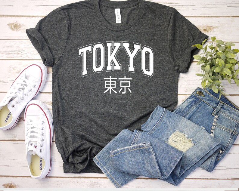 K&ouml;nnte beinhalten: Ein dunkelgraues T-Shirt mit wei&szlig;em Text, auf dem "TOKYO" und die japanischen Schriftzeichen f&uuml;r Tokio stehen. Das Shirt liegt flach auf einer Holzoberfl&auml;che mit einem Paar wei&szlig;en Turnschuhen und einer blauen Jeans.
