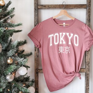 K&ouml;nnte beinhalten: Ein staubiges rosafarbenes T-Shirt mit dem Wort "TOKYO" in wei&szlig;en Blockbuchstaben und den japanischen Schriftzeichen f&uuml;r "Tokyo" darunter.
