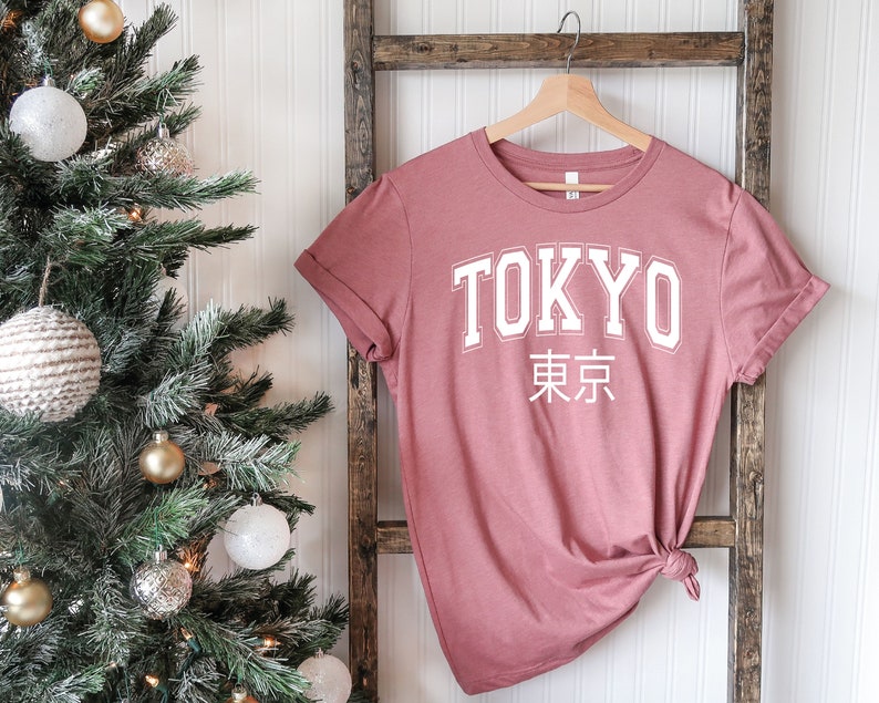 K&ouml;nnte beinhalten: Ein staubiges rosafarbenes T-Shirt mit dem Wort "TOKYO" in wei&szlig;en Blockbuchstaben und den japanischen Schriftzeichen f&uuml;r "Tokyo" darunter.
