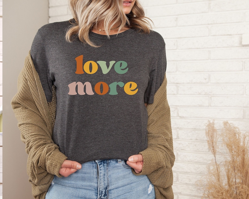 May include: Dark gray t-shirt with the words "love more" in retro-style lettering. The word "love" is in orange, yellow, and green, while "more" is in pink, orange, and green. The shirt is a casual style.