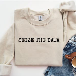 K&ouml;nnte beinhalten: Ein beiger Pullover mit dem Aufdruck "SEIZE THE DATA" in Schwarz auf der Vorderseite.