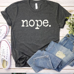 May include: A dark gray t-shirt with the word "nope" printed in white on the front. The shirt is laid out on a white wooden surface with a pair of white sneakers and a pair of blue jeans.