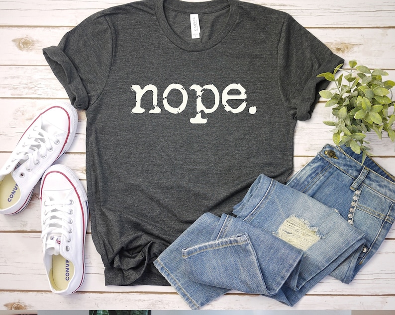 May include: A dark gray t-shirt with the word "nope" printed in white on the front. The shirt is laid out on a white wooden surface with a pair of white sneakers and a pair of blue jeans.