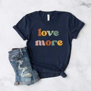 May include: Navy blue t-shirt with the words "love more" in retro-style lettering. The text is in orange, green, yellow, and pink. The shirt is paired with denim shorts.