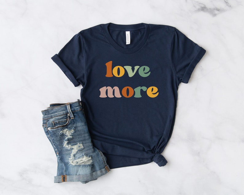 May include: Navy blue t-shirt with the words "love more" in retro-style lettering. The text is in orange, green, yellow, and pink. The shirt is paired with denim shorts.