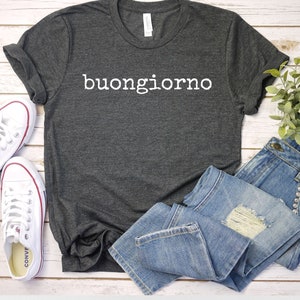 May include: A dark gray t-shirt with the Italian phrase "buongiorno" printed in white on the front. The shirt is laid out on a white wooden surface with a pair of white sneakers and a pair of blue jeans.