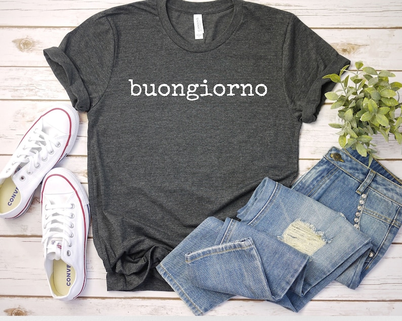 May include: A dark gray t-shirt with the Italian phrase "buongiorno" printed in white on the front. The shirt is laid out on a white wooden surface with a pair of white sneakers and a pair of blue jeans.