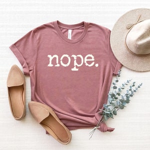 May include: A mauve t-shirt with the word "nope" printed in white on the front. The shirt is laid flat on a white surface with a hat and some greenery.
