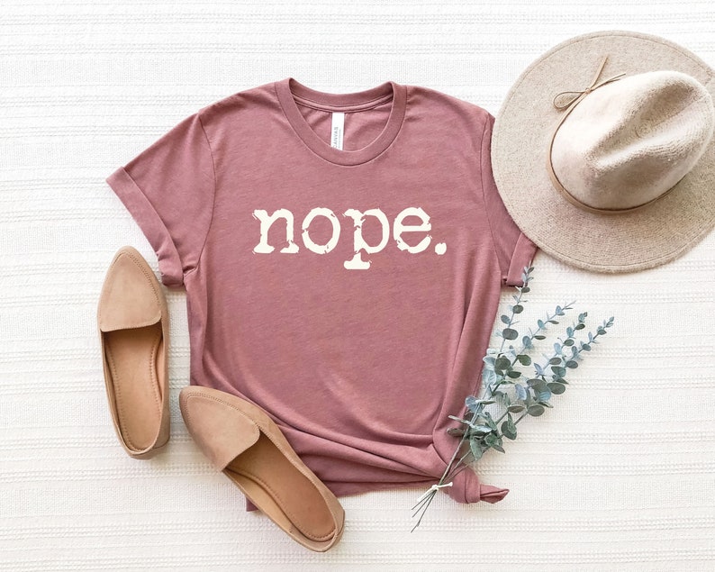 May include: A mauve t-shirt with the word "nope" printed in white on the front. The shirt is laid flat on a white surface with a hat and some greenery.