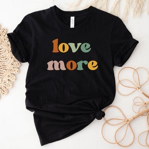 May include: Black t-shirt with the words "love more" in retro-style lettering. The text is in shades of orange, green, pink, and yellow. The shirt is made of a soft material and is likely a unisex tee.