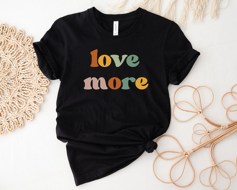 May include: Black t-shirt with the words "love more" in retro-style lettering. The text is in shades of orange, green, pink, and yellow. The shirt is made of a soft material and is likely a unisex tee.